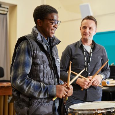 A boy in a blue checked t-shirt and camouflaged gilet plays the drums. He is stood next to a man in a grey shirt. Both are smiling.